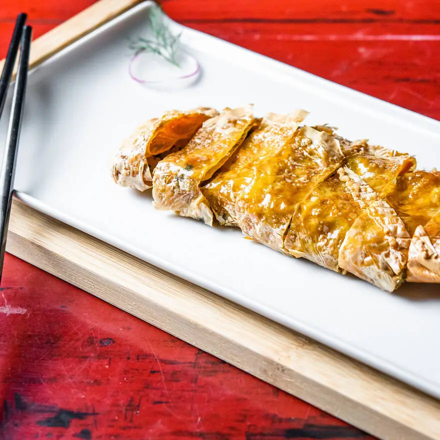 Sliced glazed pastry-wrapped roll in amber sauce on a white rectangular plate with black chopsticks, on a red lacquered table