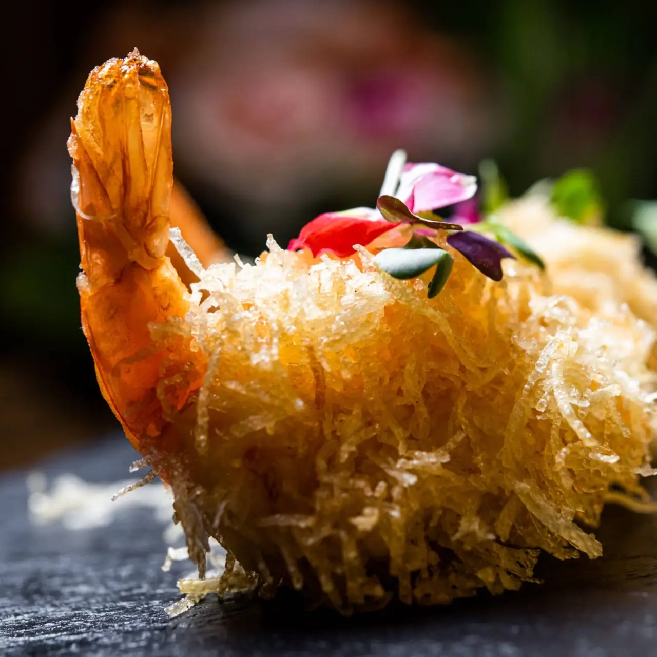 Close-up of a crispy shredded-pastry-coated fried prawn topped with edible flower petals and microgreens on a dark surface