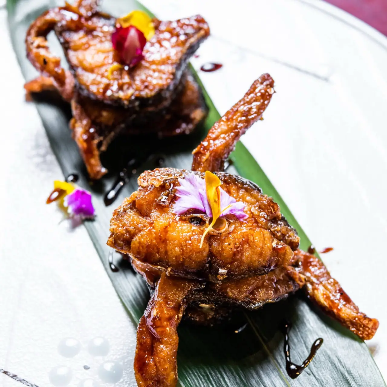 Two portions of caramel-glazed fried meat garnished with pink and yellow edible flowers on a green leaf over a white plate