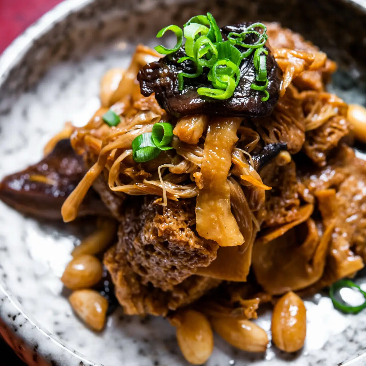 Braised mixed mushrooms and honeycomb tripe in dark sauce topped with sliced spring onion and garlic cloves in a speckled bowl
