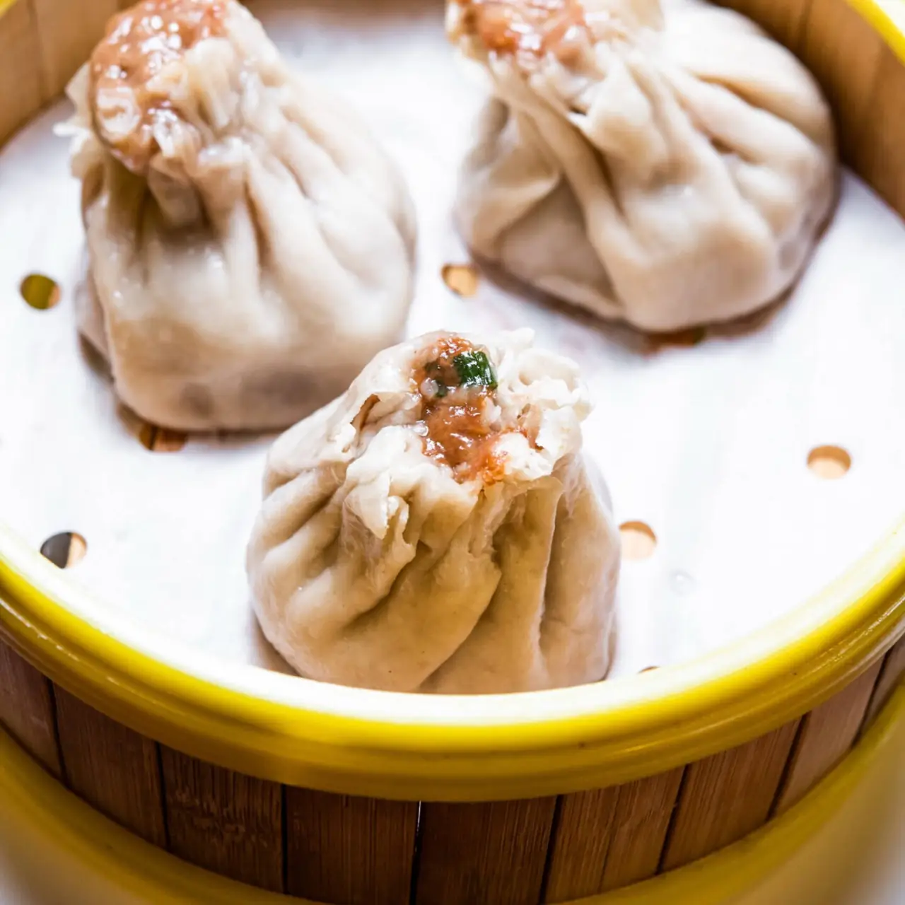 Three steamed soup dumplings with pleated tops in a yellow-rimmed bamboo steamer basket