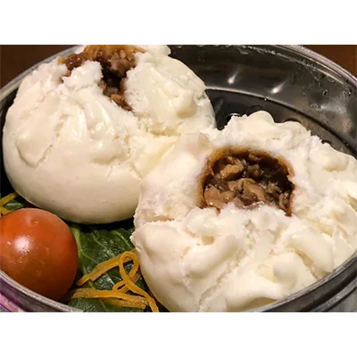 Two steamed pork baozi split open in a metal steamer basket, a cherry tomato and shredded carrot beside them