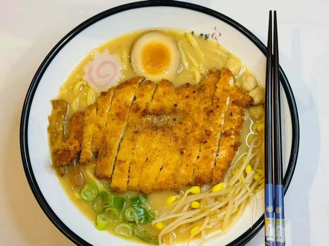A bowl of ramen in creamy broth topped with a sliced breaded cutlet, a halved egg, bean sprouts and chopsticks
