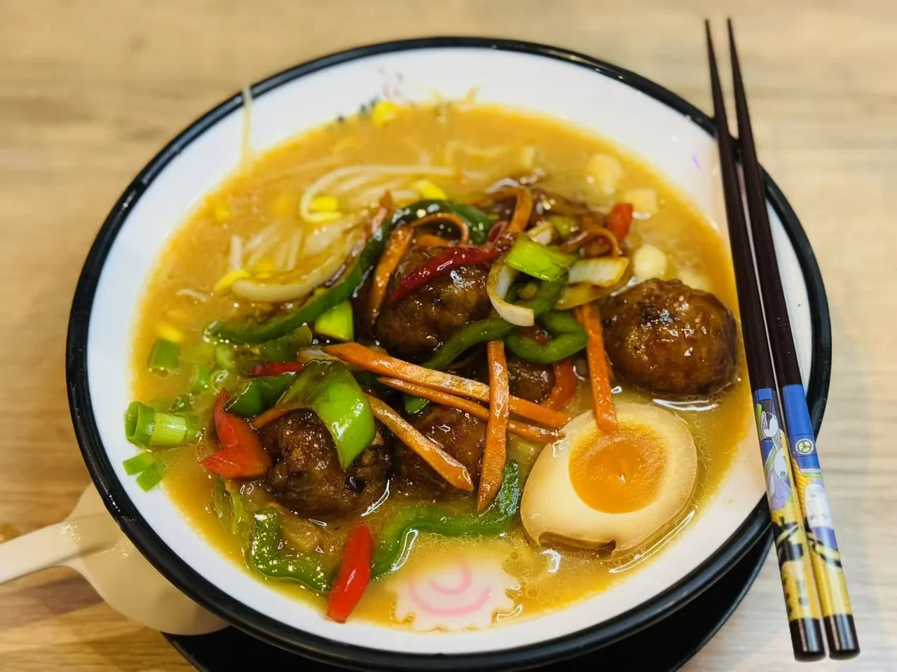 A bowl of ramen in golden broth with meatballs, stir-fried peppers and carrot, a halved egg and chopsticks
