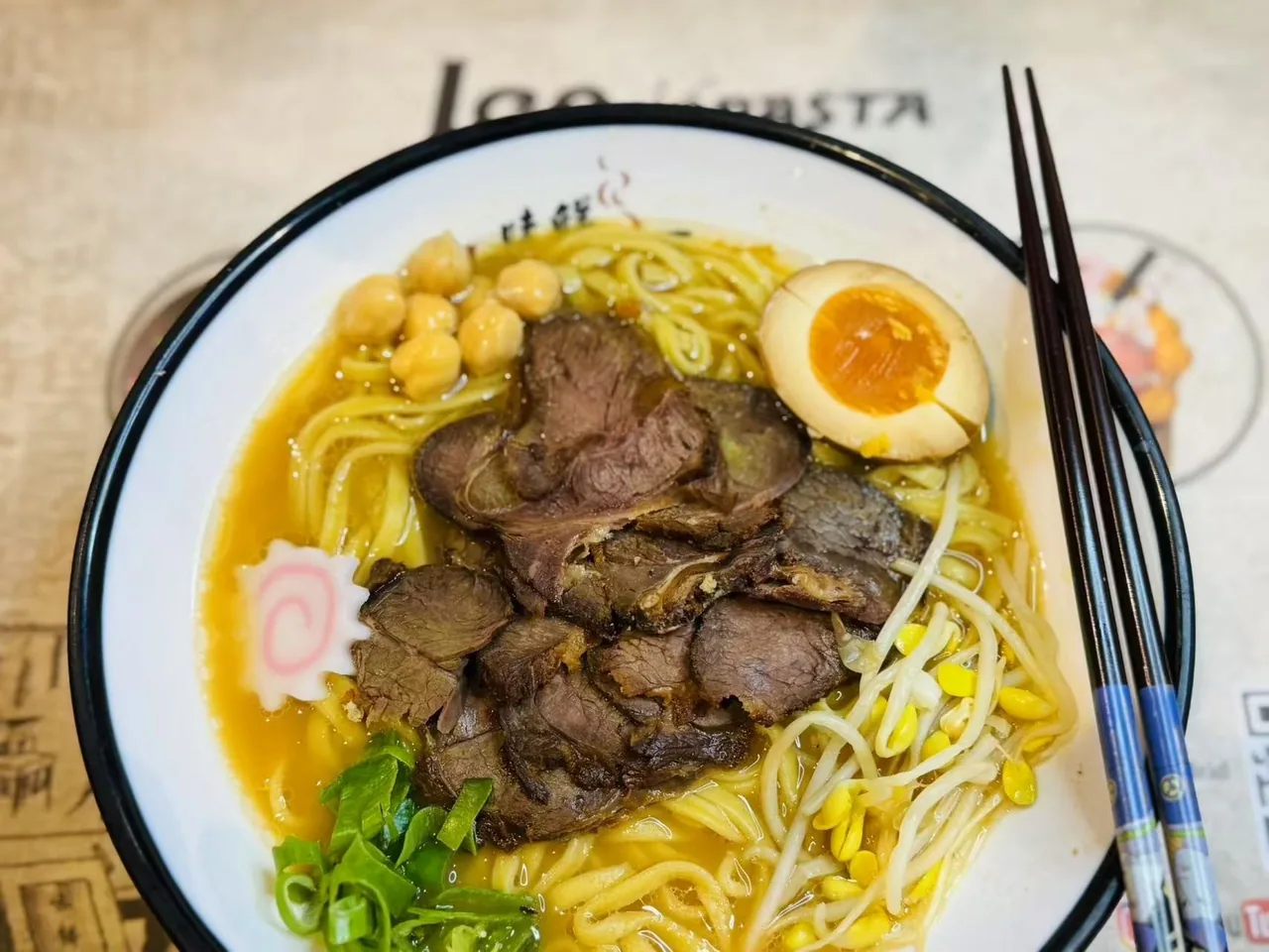 A bowl of ramen with slices of braised beef, chickpeas, a halved egg and bean sprouts beside chopsticks