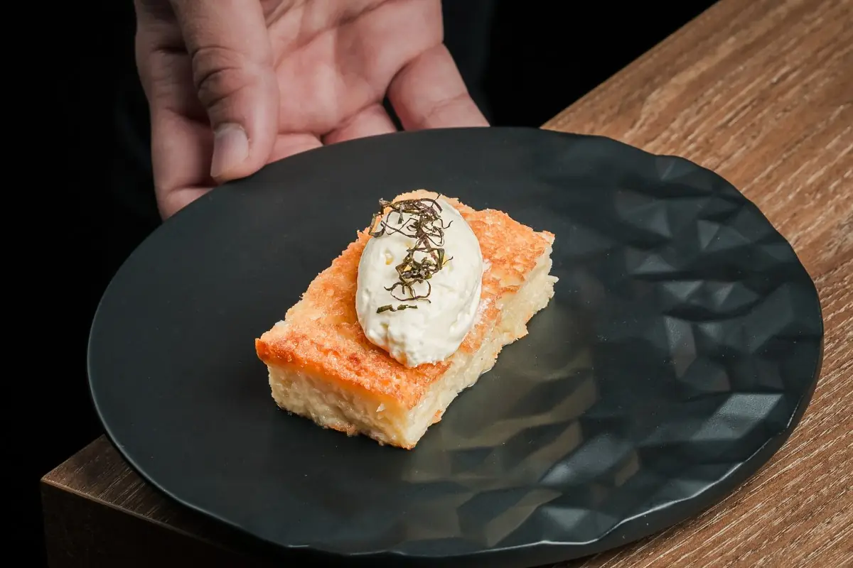 Hand presenting a black faceted plate holding a golden-crusted milk-soaked cake square topped with a cream quenelle and dark shredded garnish.
