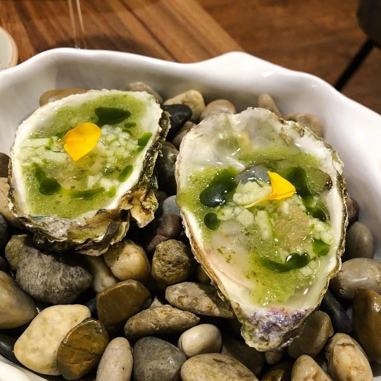Two oysters topped with green sauce and a yellow piece in a bowl of dark stones