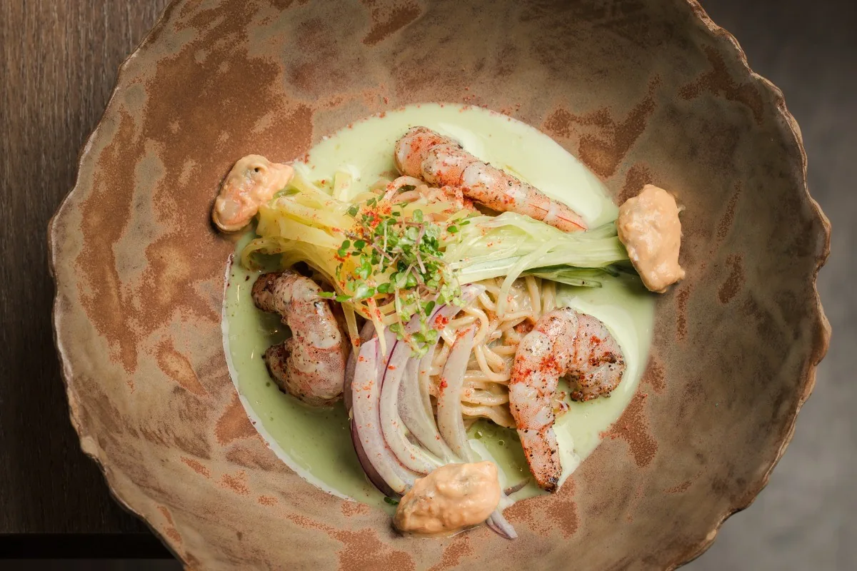 A round bowl with noodles, pieces of prawn and thin slices of onion