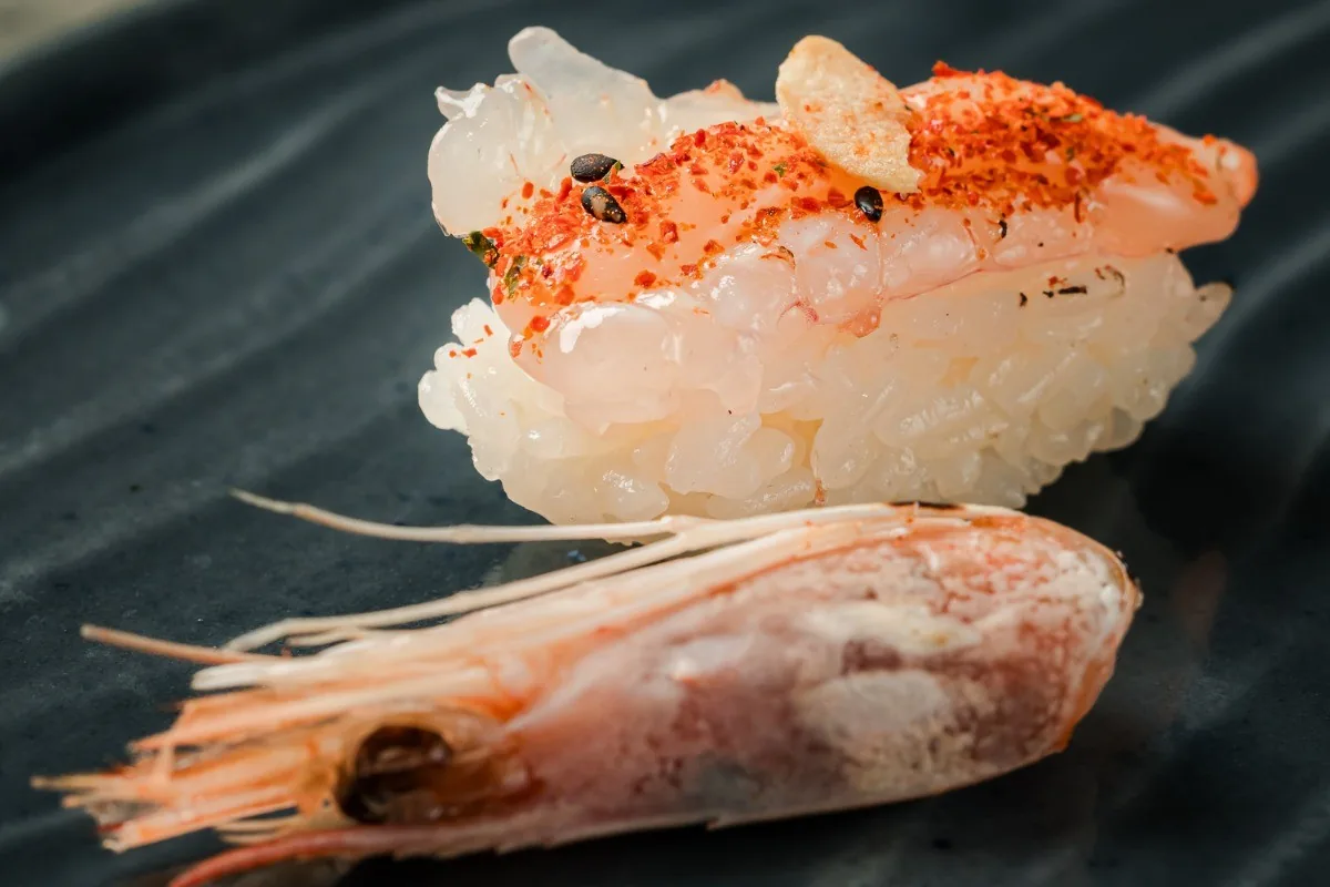 A nigiri topped with orange-spiced fish next to a raw prawn on a dark plate