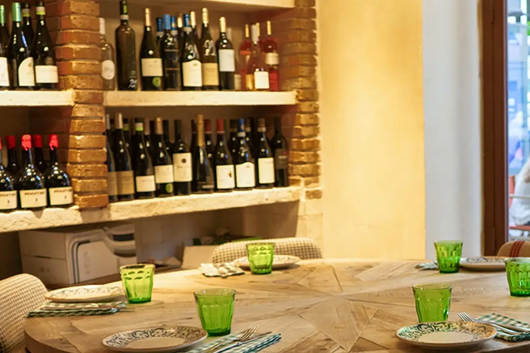 Restaurant interior with a round wooden table set with green glass tumblers and patterned plates, backed by a brick-framed wall of wine bottles