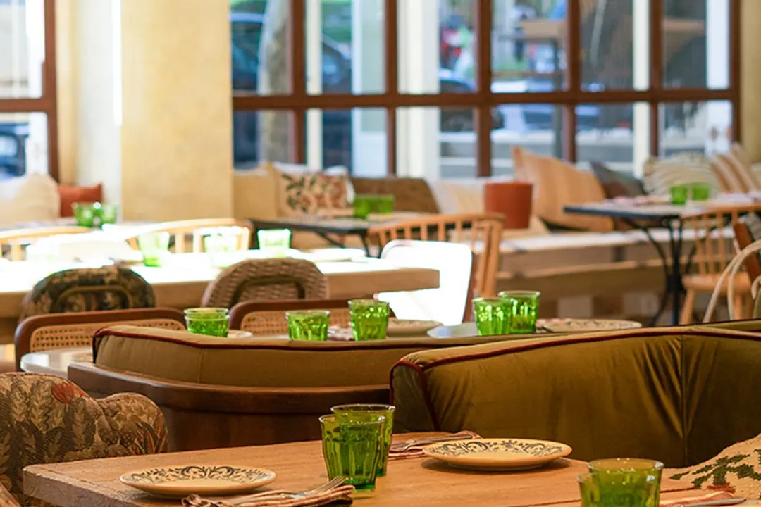 Dining room with cushioned banquettes, wooden tables set with green glasses and patterned plates, large windows behind