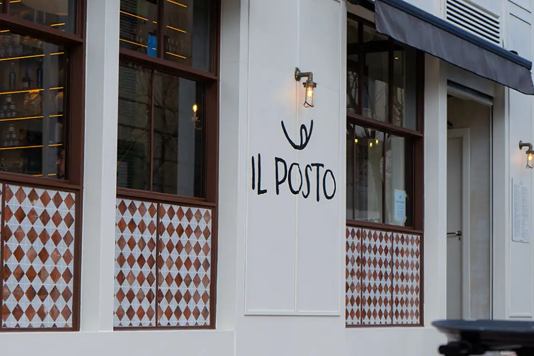 Exterior storefront of Il Posto with white facade, diamond-tiled base, wall sconce and hand-painted IL POSTO sign