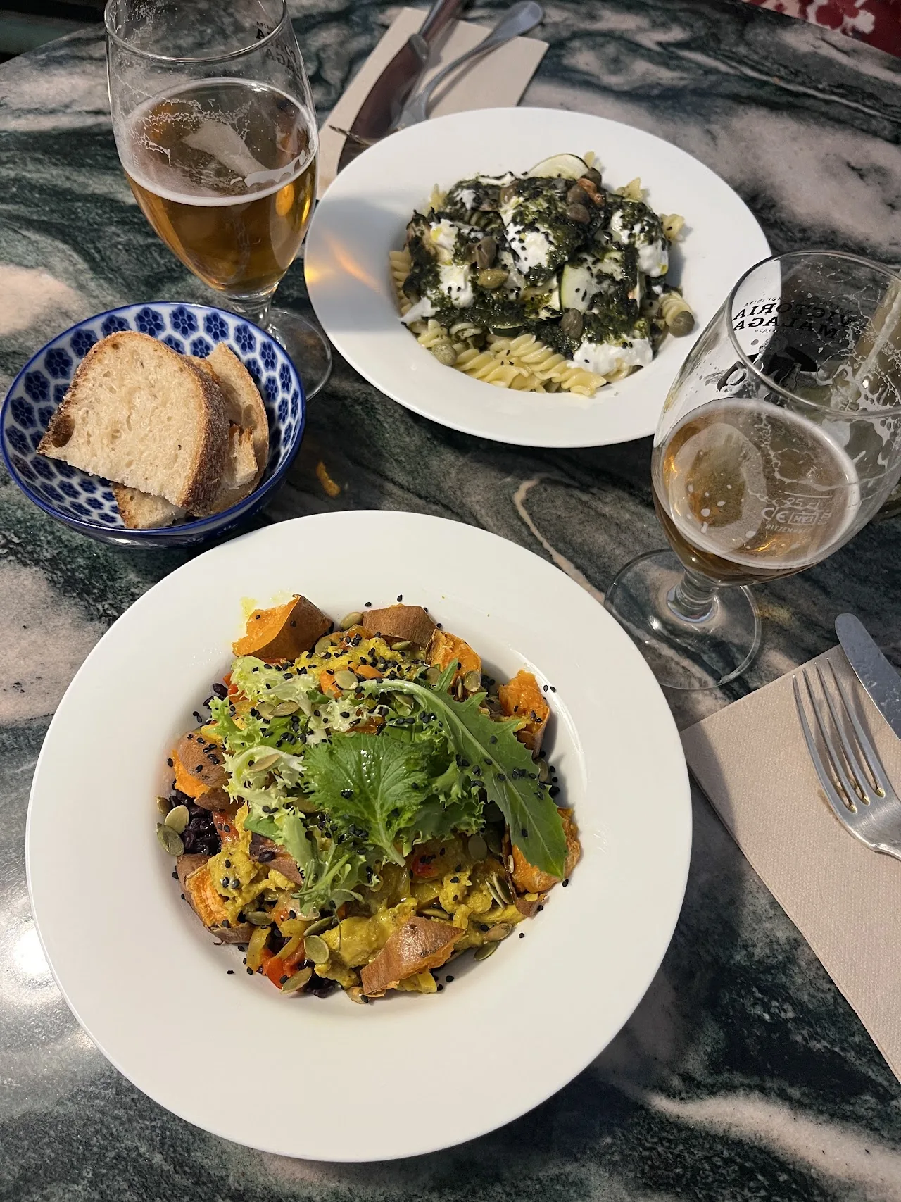 An overhead view of a green pasta dish and a mixed salad on a table with drinks