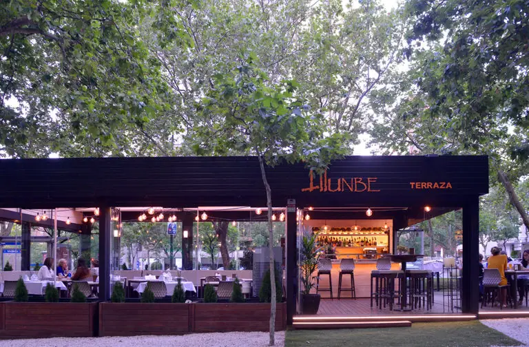 Restaurant exterior at dusk under leafy trees, black-paneled facade lit with string lights and a sign reading ILLUNBE TERRAZA above an open bar area with stools.