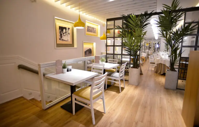 Restaurant interior dining room with white tables and chairs, framed photographs on the wall, potted palm plants and wood flooring.