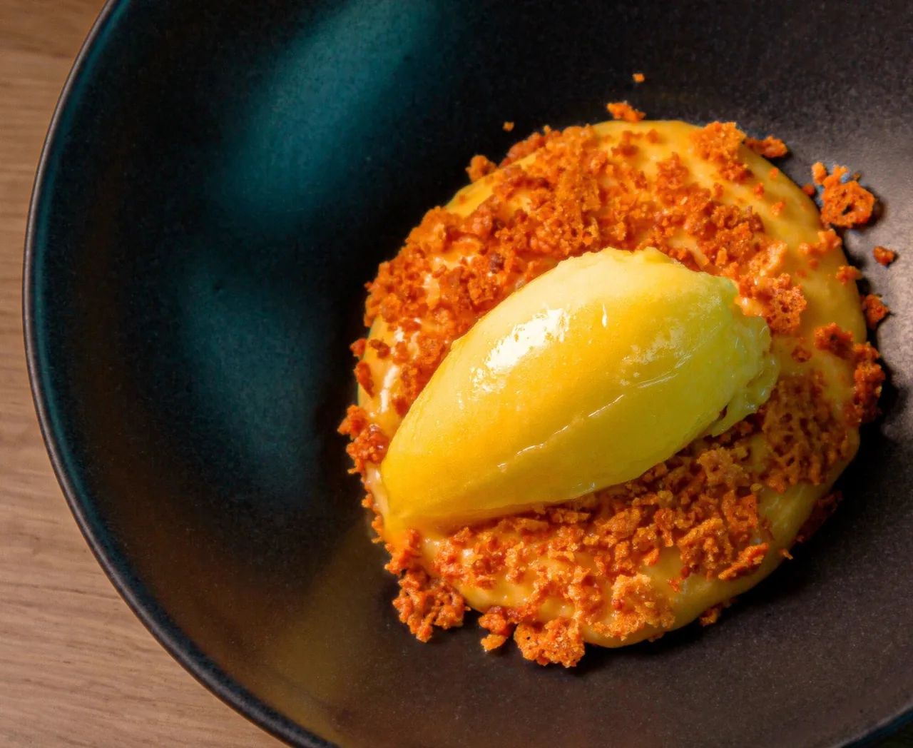 A dark bowl with a scoop of yellow sorbet on orange crumbs