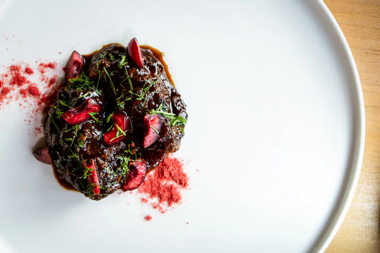Braised meat dish topped with fresh herbs, sliced cherries, and a red spice powder