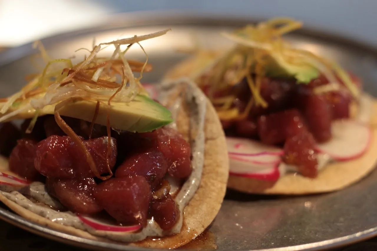 Two small tortillas topped with diced raw tuna, avocado and crispy fried strands on a metal plate