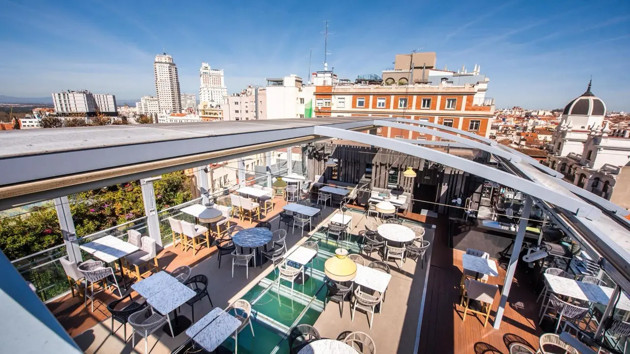 Rooftop terrace with marble tables and rattan chairs under a retractable glass canopy, overlooking the Madrid skyline including Torre de Madrid.