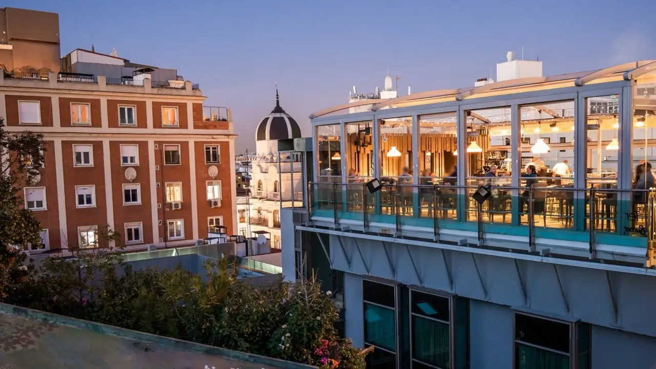 Glass-walled rooftop terrace bar at dusk with patrons seated at tables, Madrid rooftops and a domed building visible in the background.
