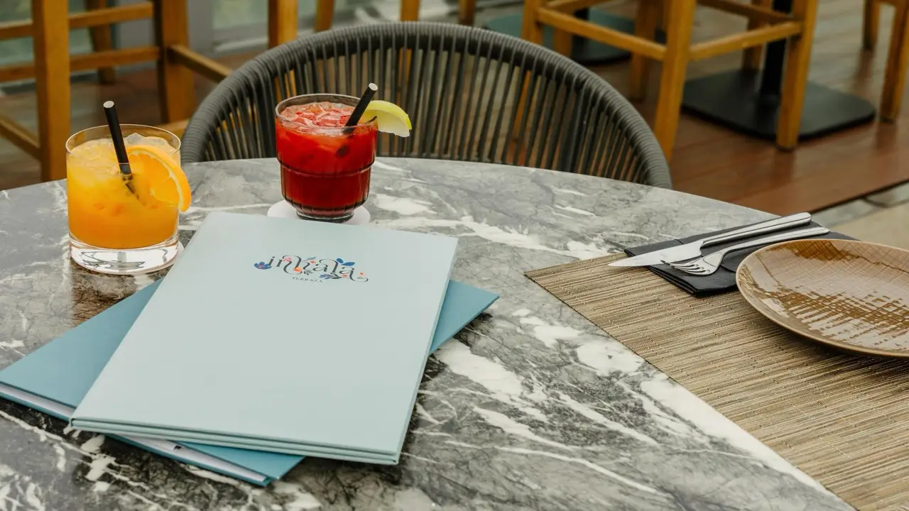 Two poured cocktails, one orange and one red, beside a table setting and a menu booklet reading Inhala Terraza on a marble table.