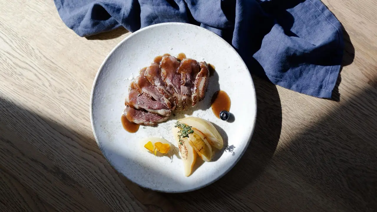Sliced roasted duck breast with pan sauce, a caramelized pear wedge with thyme, and a blueberry, plated on a white ceramic dish.