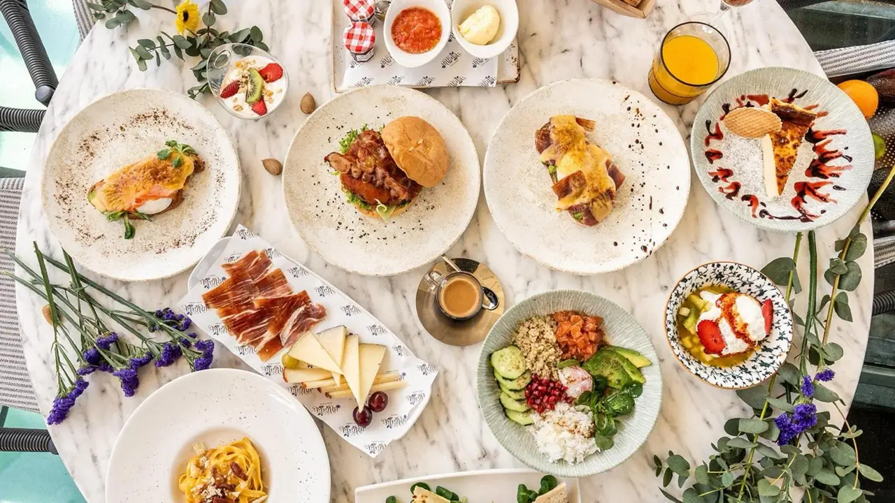 Overhead view of a brunch spread on a marble table including eggs benedict, a burger, jamon and cheese board, a poke bowl, coffee, orange juice and dessert plates.