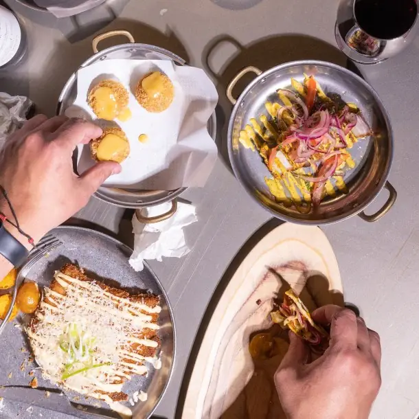 Overhead view of plated dishes on a table, hands picking up fried bites and tacos, glass of red wine
