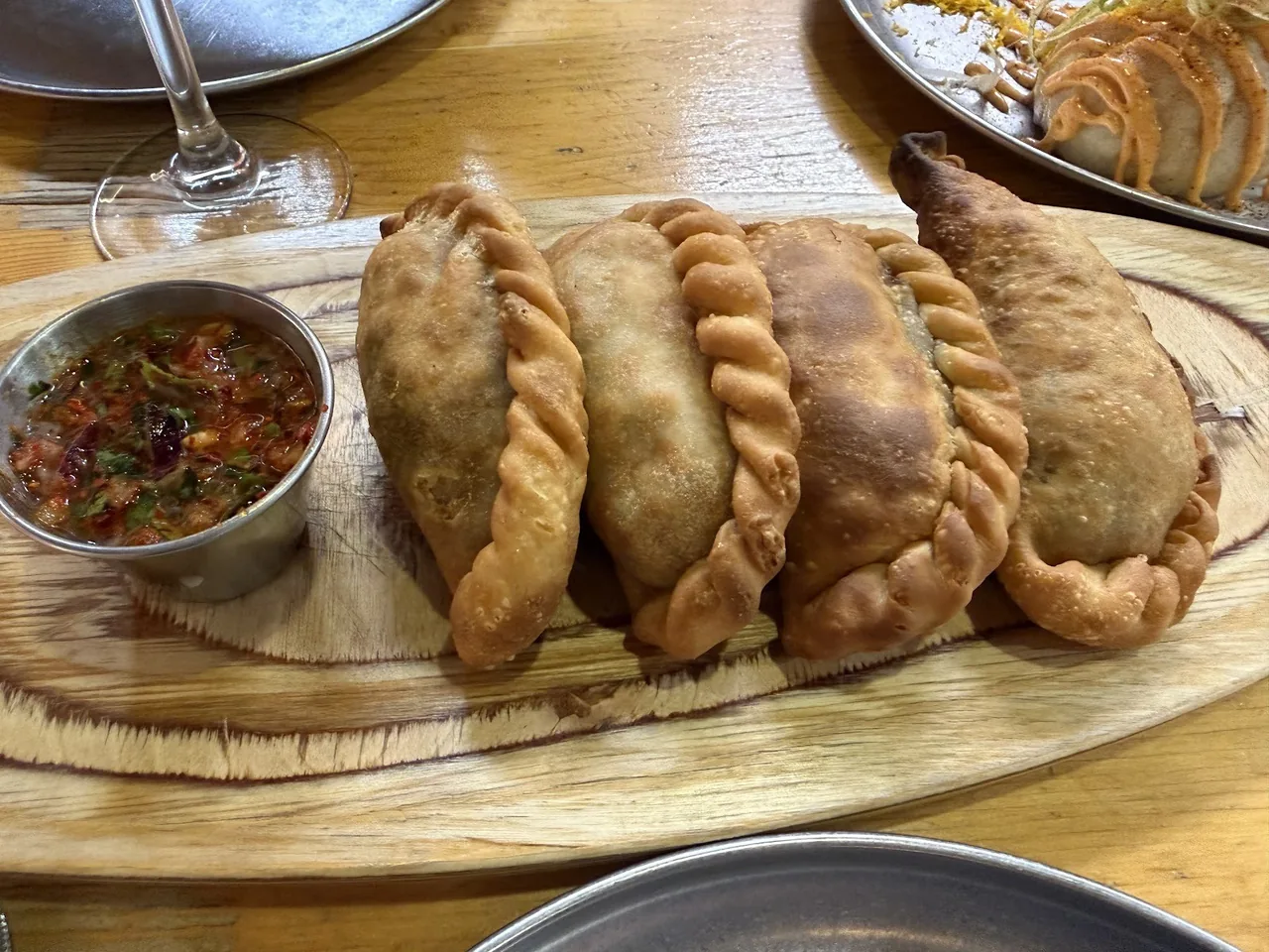 A row of golden empanadas on a wooden board with a small dish of red salsa
