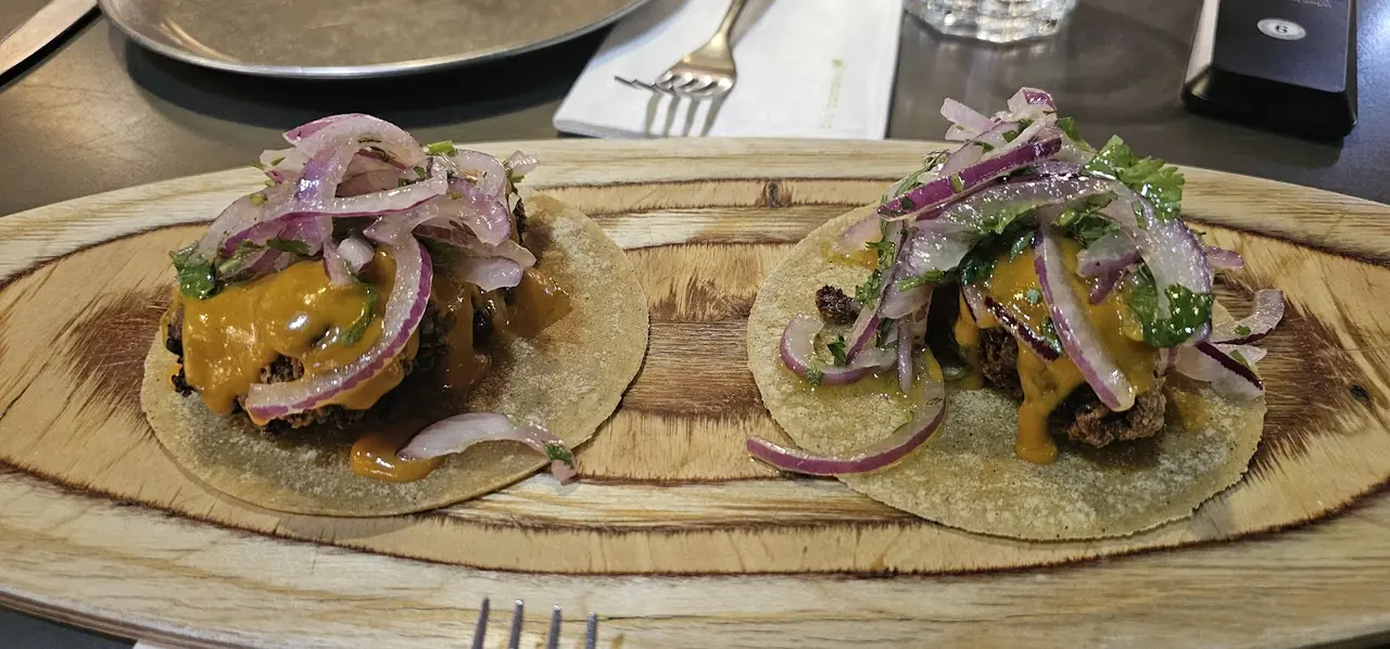 Two tacos on a wooden board topped with red onion, cilantro and orange sauce