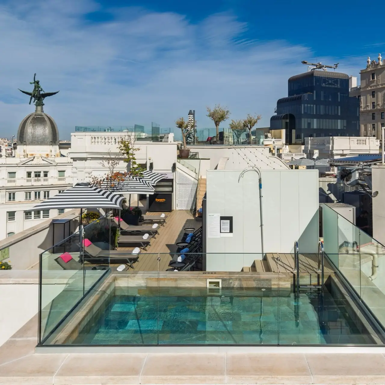 Rooftop terrace with a small plunge pool, sun loungers and striped umbrellas overlooking Madrid rooftops and a domed building topped with a winged statue