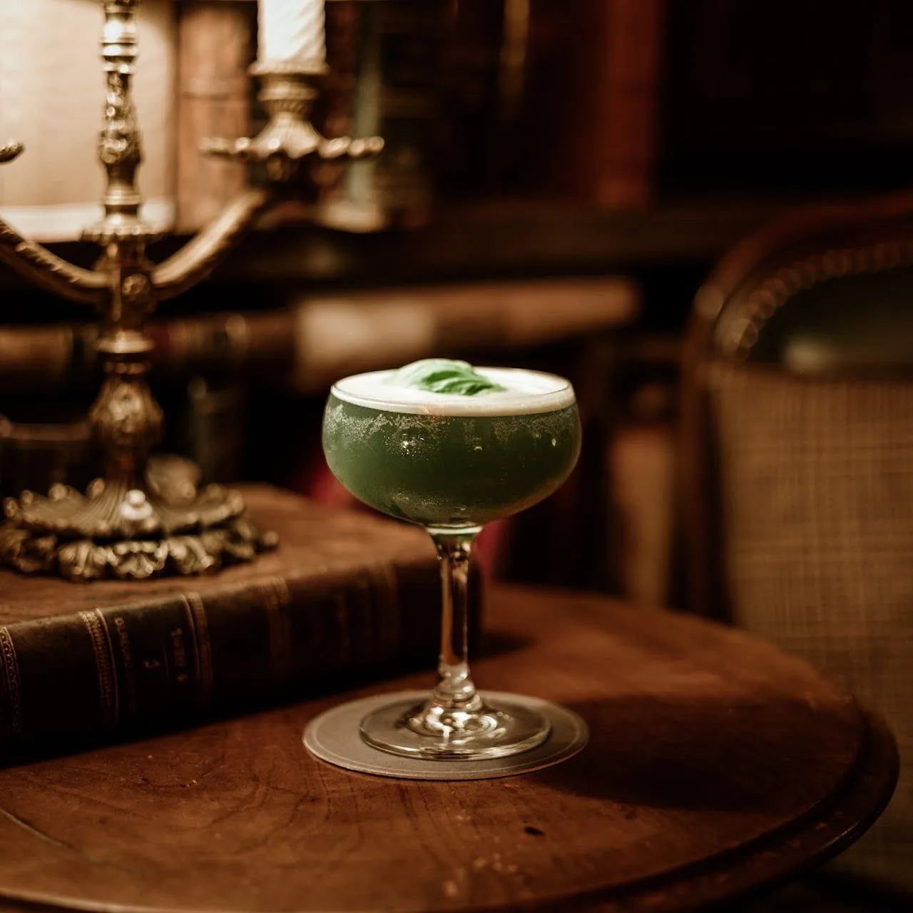 A green frothy cocktail in a coupe glass on a round wooden table with an old book behind it