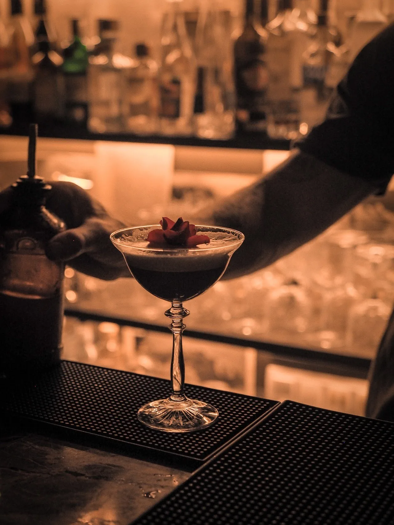 A cocktail in a coupe glass on a bar mat with a bartender's arm behind it