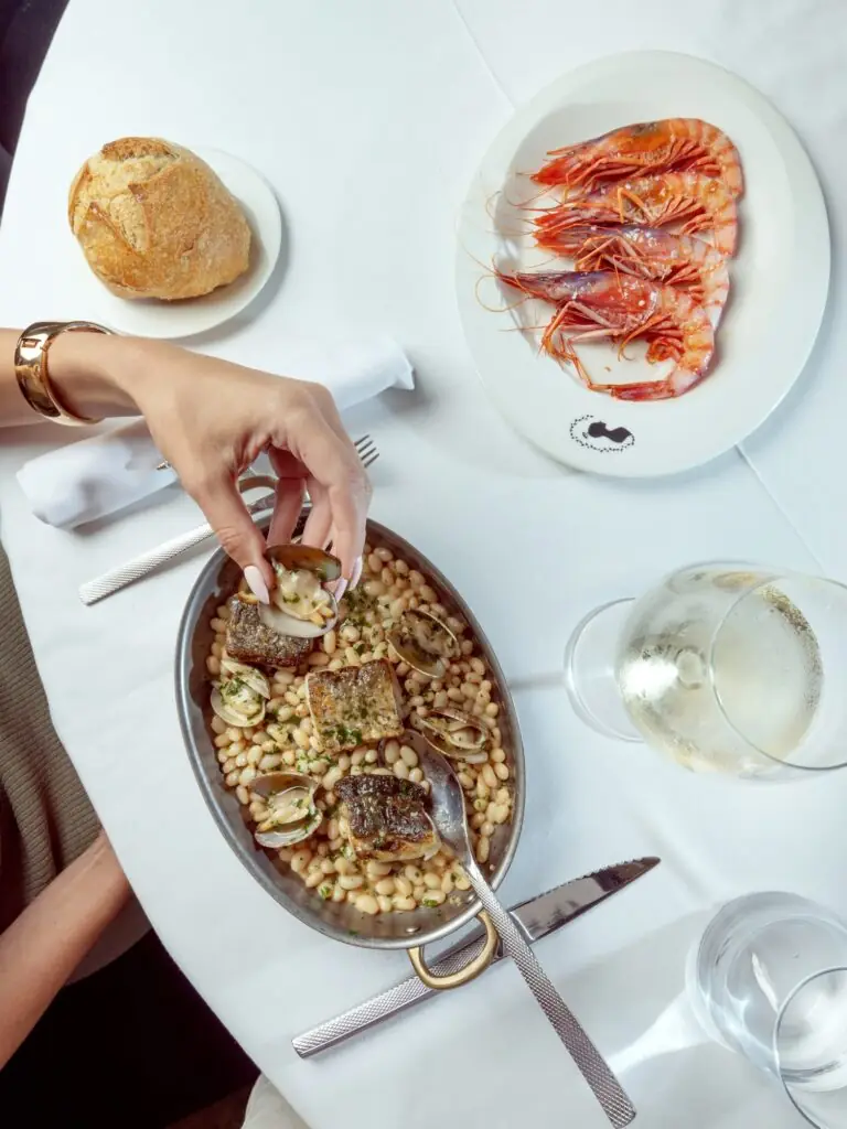 Overhead shot of a pan of pan-seared fish fillets with white beans and clams, alongside a plate of whole grilled red prawns and a bread roll, at Jacqueline Barcelona