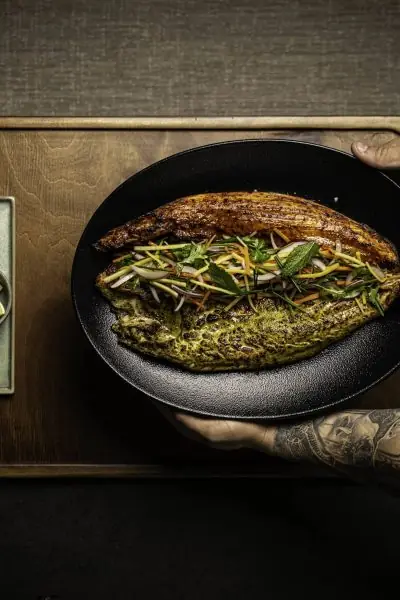 Whole grilled fish topped with fresh herbs and julienned vegetables, served on a black plate at Jaiba MX