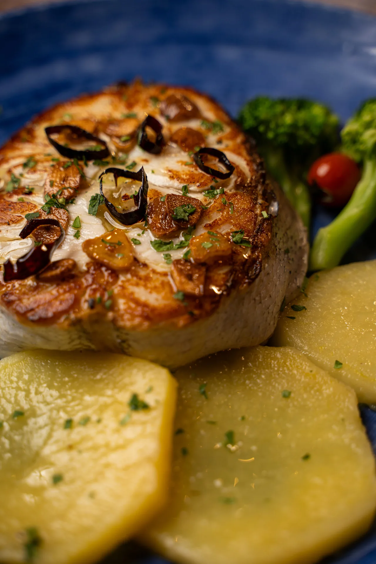 A thick seared fish steak topped with sliced garlic, dried chilli rings and parsley on a blue plate, with sliced boiled potatoes, broccoli and a cherry tomato