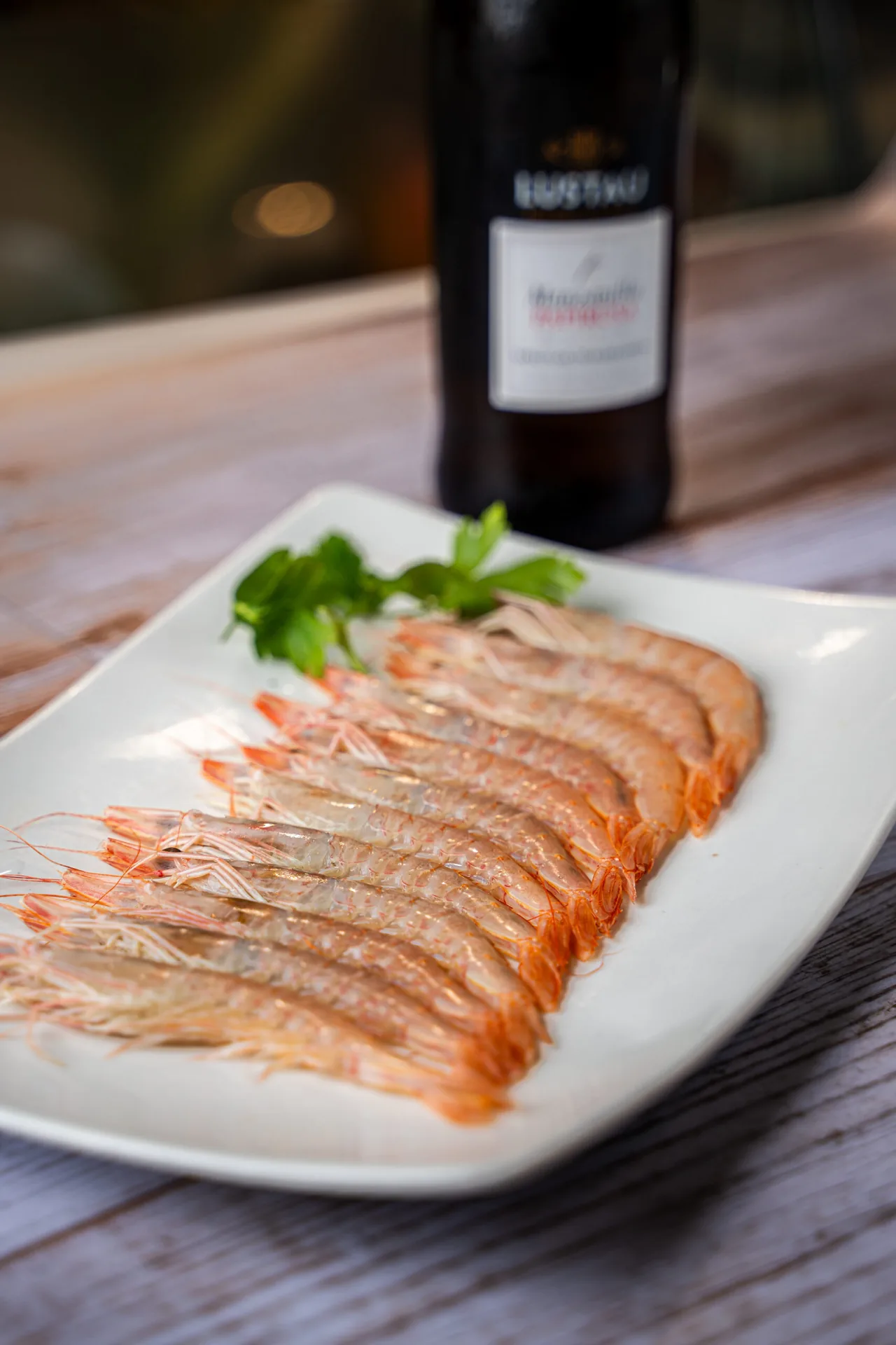 A row of raw langoustines laid head-to-tail on a white rectangular plate with a sprig of parsley, a dark bottle of manzanilla behind on a weathered wooden table