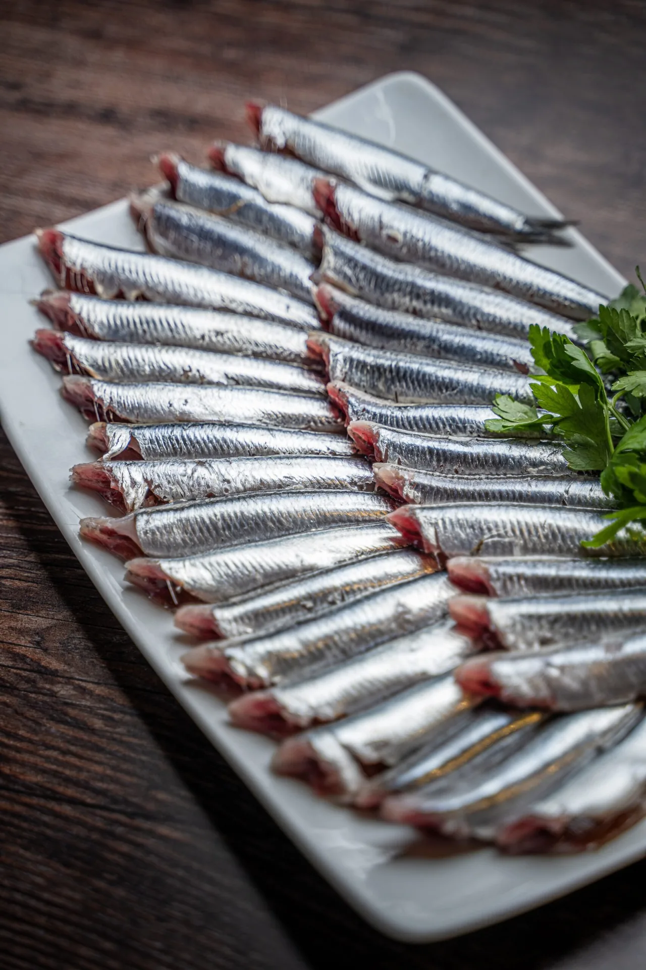 Rows of fresh anchovy fillets on a white rectangular plate with parsley