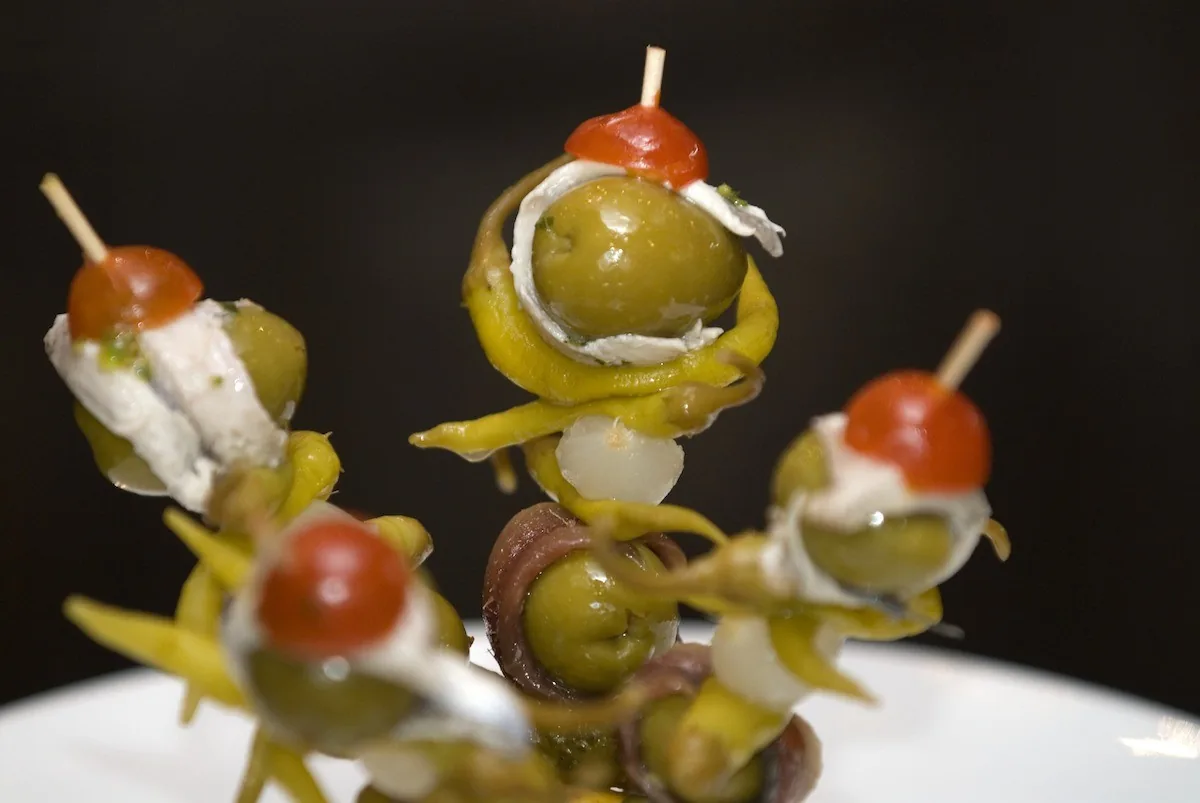 Skewers of green pickled peppers, olives and anchovy on a white plate