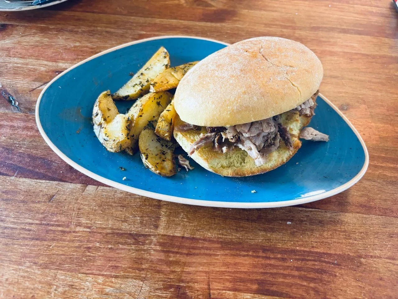 A round roll filled with shredded roast meat on a blue plate next to herbed potato wedges