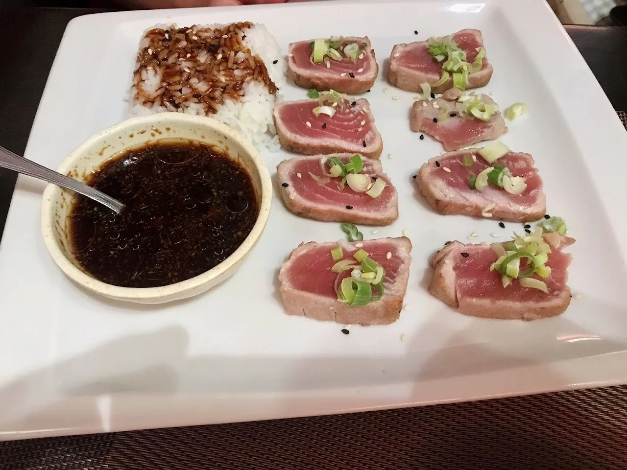 Slices of seared fish with a green garnish on a white square plate beside a bowl of dark sauce