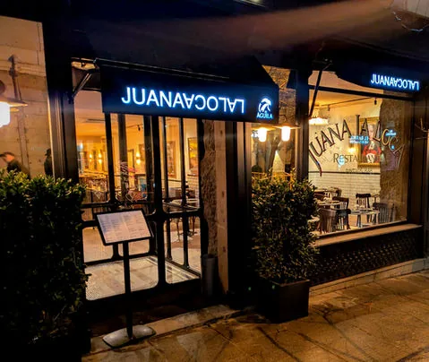 Street frontage at night: a lit blue sign over the glass front, a menu board on the pavement and clipped bay trees either side of the door