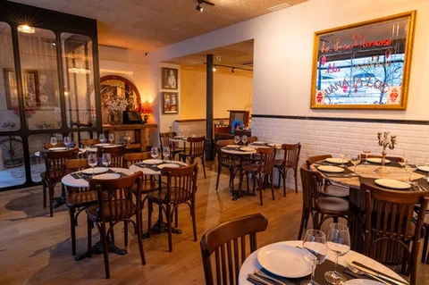 Dining room with bentwood chairs around small round tables laid with white cloths, white brick walls, a framed picture and a wine cabinet at the far end