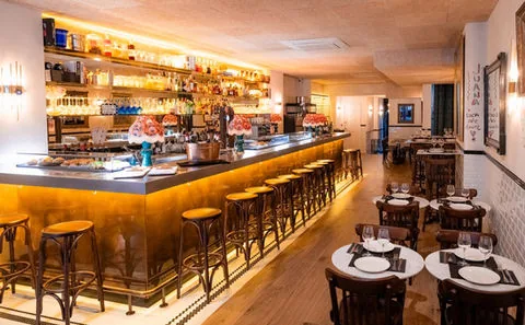 A long brass-fronted bar with wooden stools along it, pintxos laid out on the counter and bottles on shelves behind, tables to the right