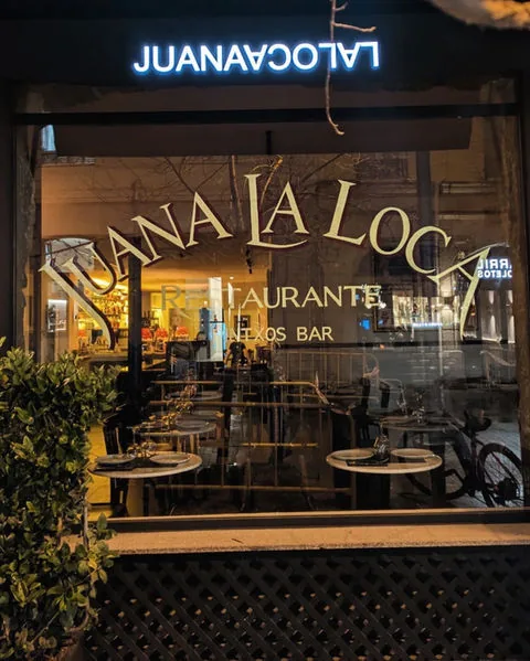 The restaurant window at night with 'Juana La Loca Restaurante, Pintxos Bar' in gold lettering across the glass, laid tables visible inside and an illuminated sign above