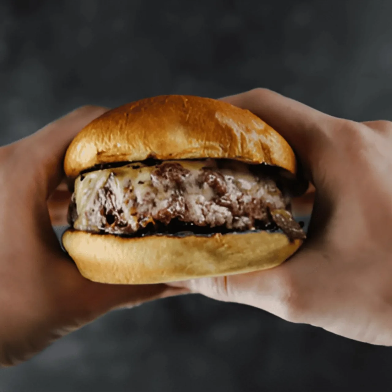 A hand holding a burger with a thick patty on a glossy bun against a dark background