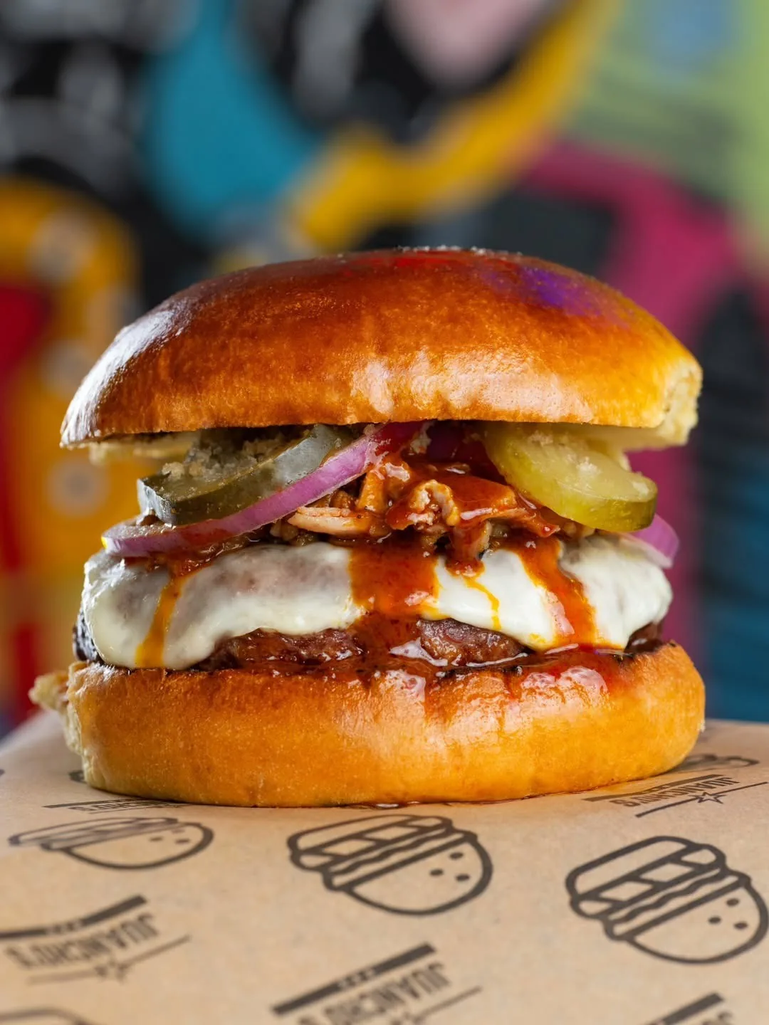 A tall burger with onion, pickles, cheese and sauce on printed paper