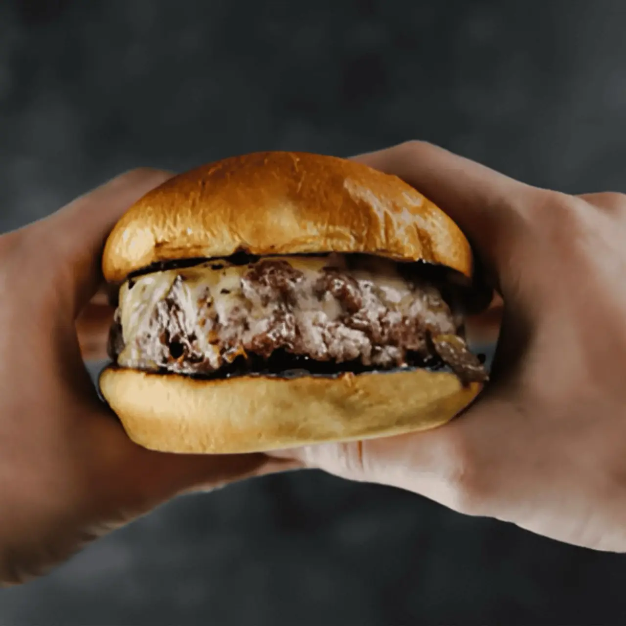 Hands holding a cheeseburger with melted cheese on a glossy brioche bun