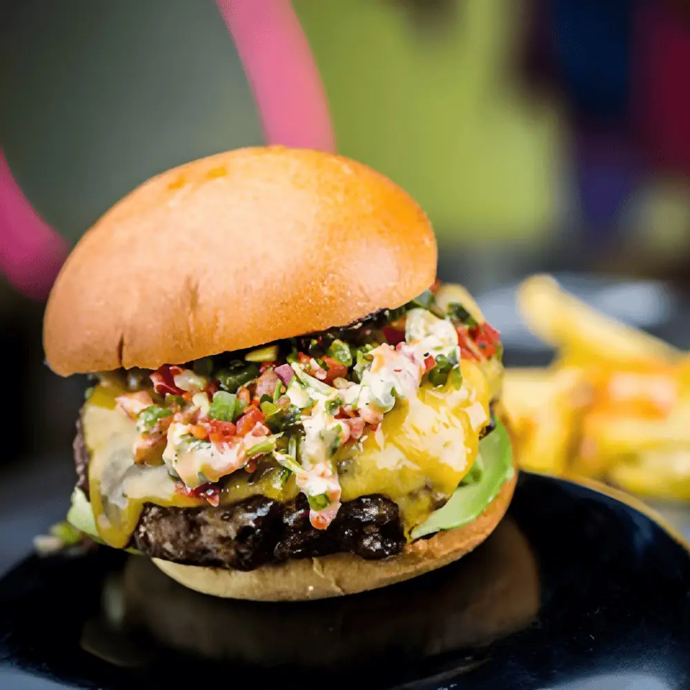 Loaded cheeseburger topped with pico de gallo and slaw on a black plate with fries behind