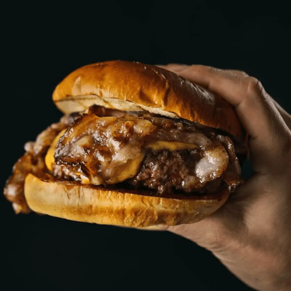 Hand holding a cheeseburger with melted cheese and caramelized onions against a dark background